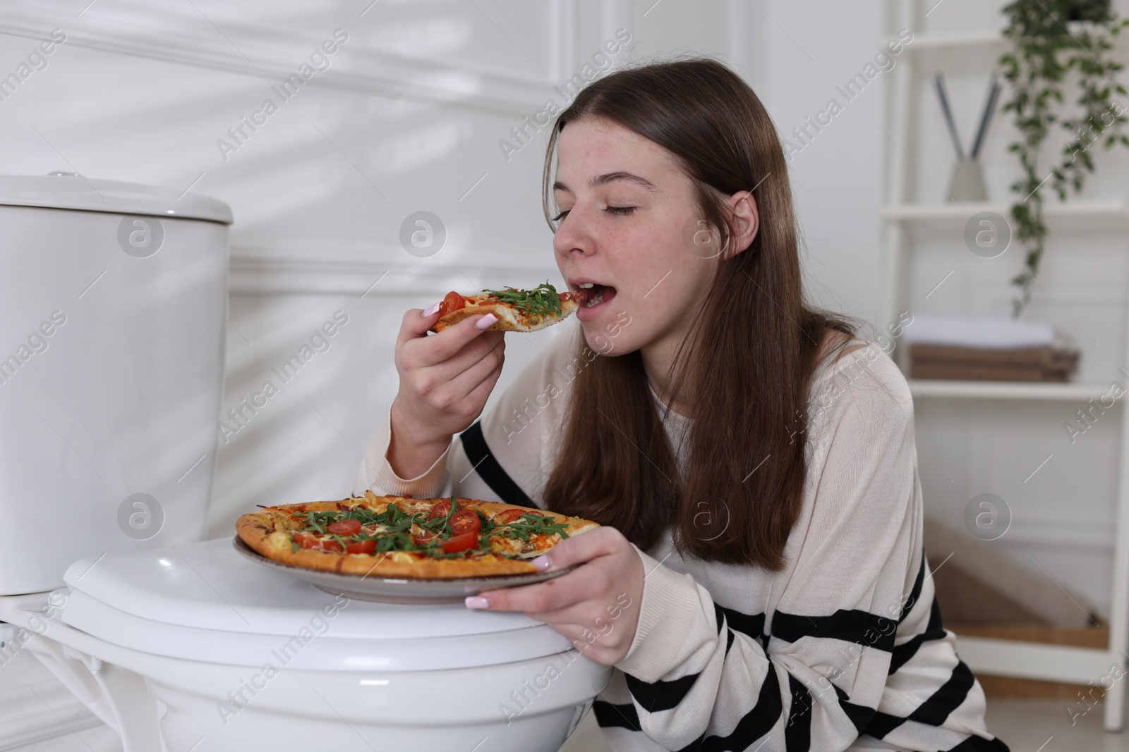 Photo of Teen girl eating pizza near toilet bowl at home. Bulimia