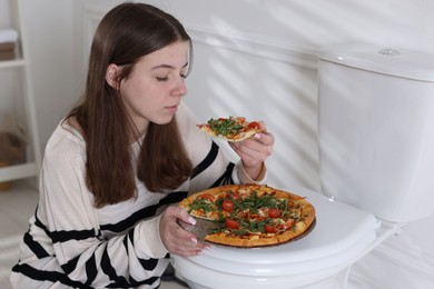 Photo of Teen girl eating pizza near toilet bowl at home. Bulimia