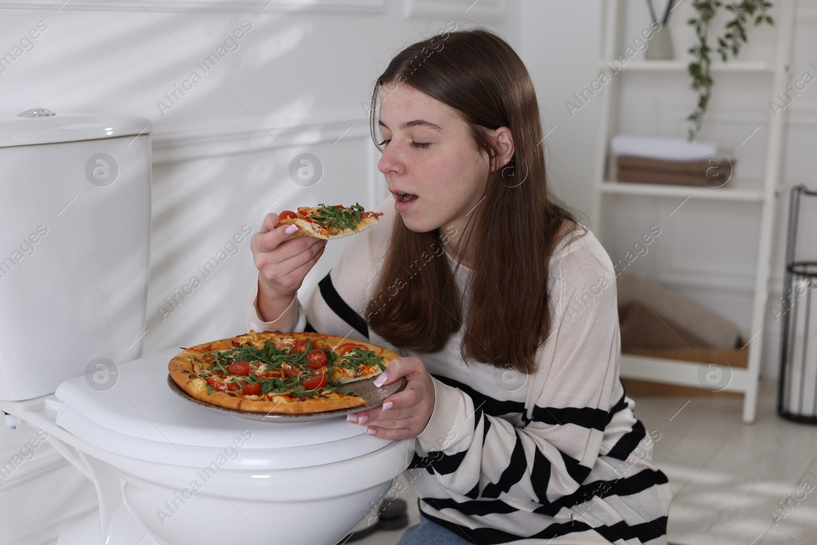 Photo of Teen girl eating pizza near toilet bowl at home. Bulimia