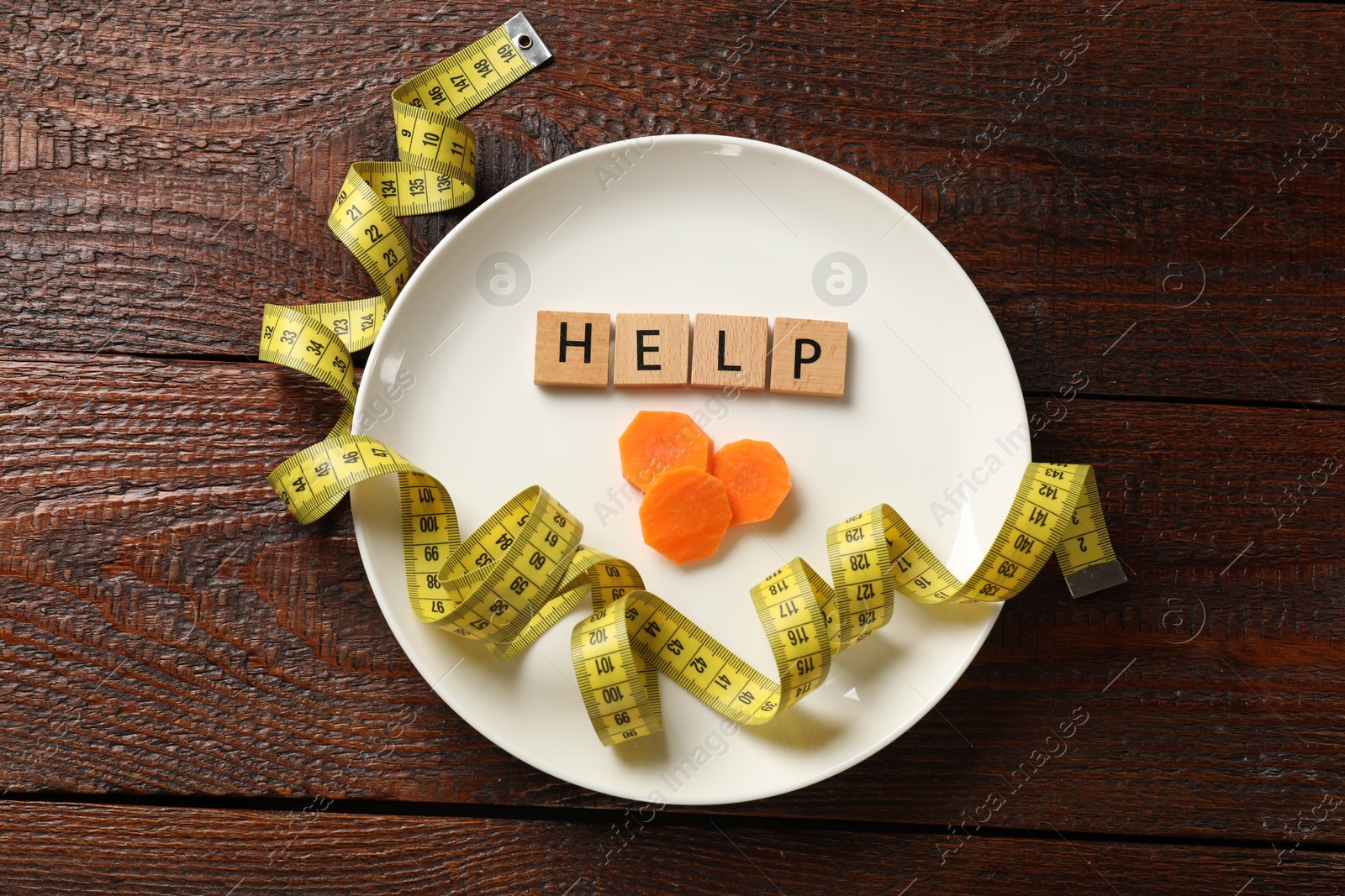 Photo of Bulimia. Squares with word Help, plate, cut carrot and measuring tape on wooden table, top view