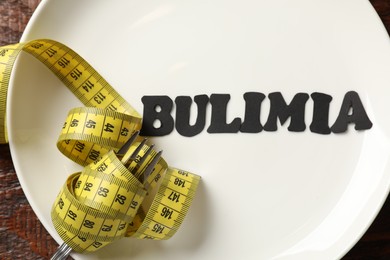 Photo of Plate with word Bulimia, fork and measuring tape on wooden table, top view