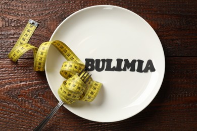 Photo of Plate with word Bulimia, fork and measuring tape on wooden table, top view