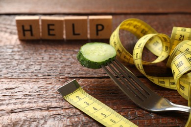 Photo of Bulimia. Squares with word Help, fork, slice of cucumber and measuring tape on wooden table, closeup