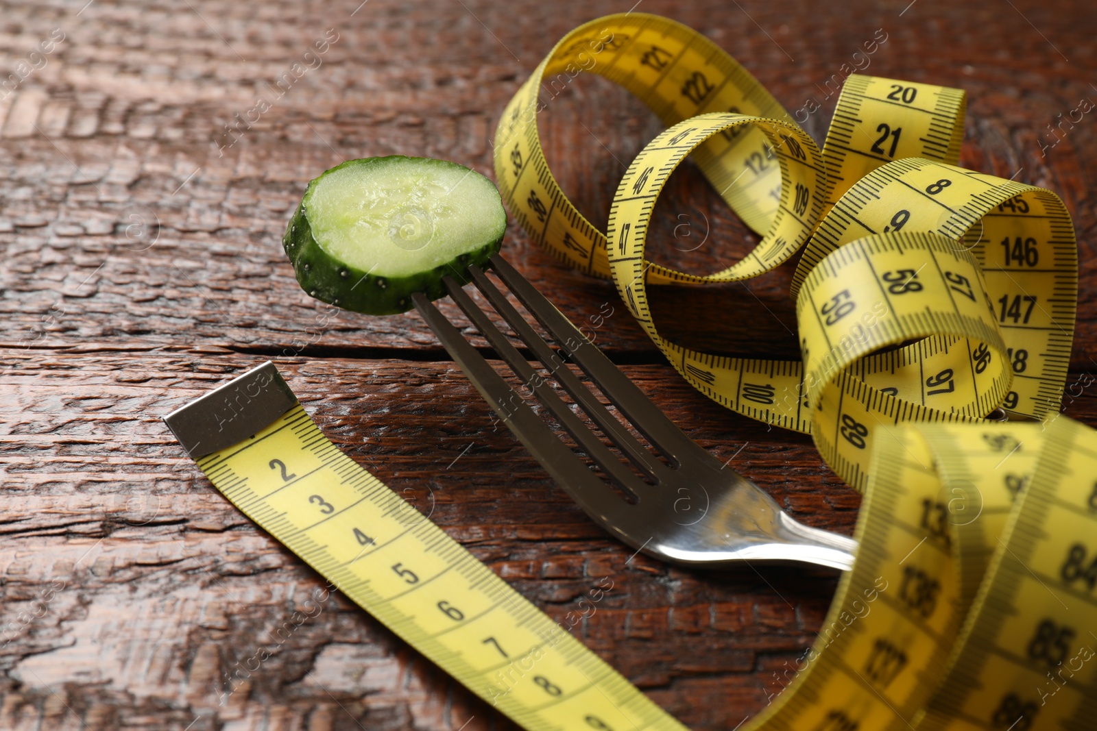 Photo of Bulimia. Fork with slice of cucumber and measuring tape on wooden table, closeup