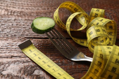 Photo of Bulimia. Fork with slice of cucumber and measuring tape on wooden table, closeup