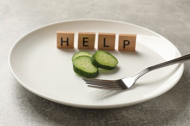 Photo of Bulimia. Squares with word Help, plate, cut cucumber and fork on grey table, closeup