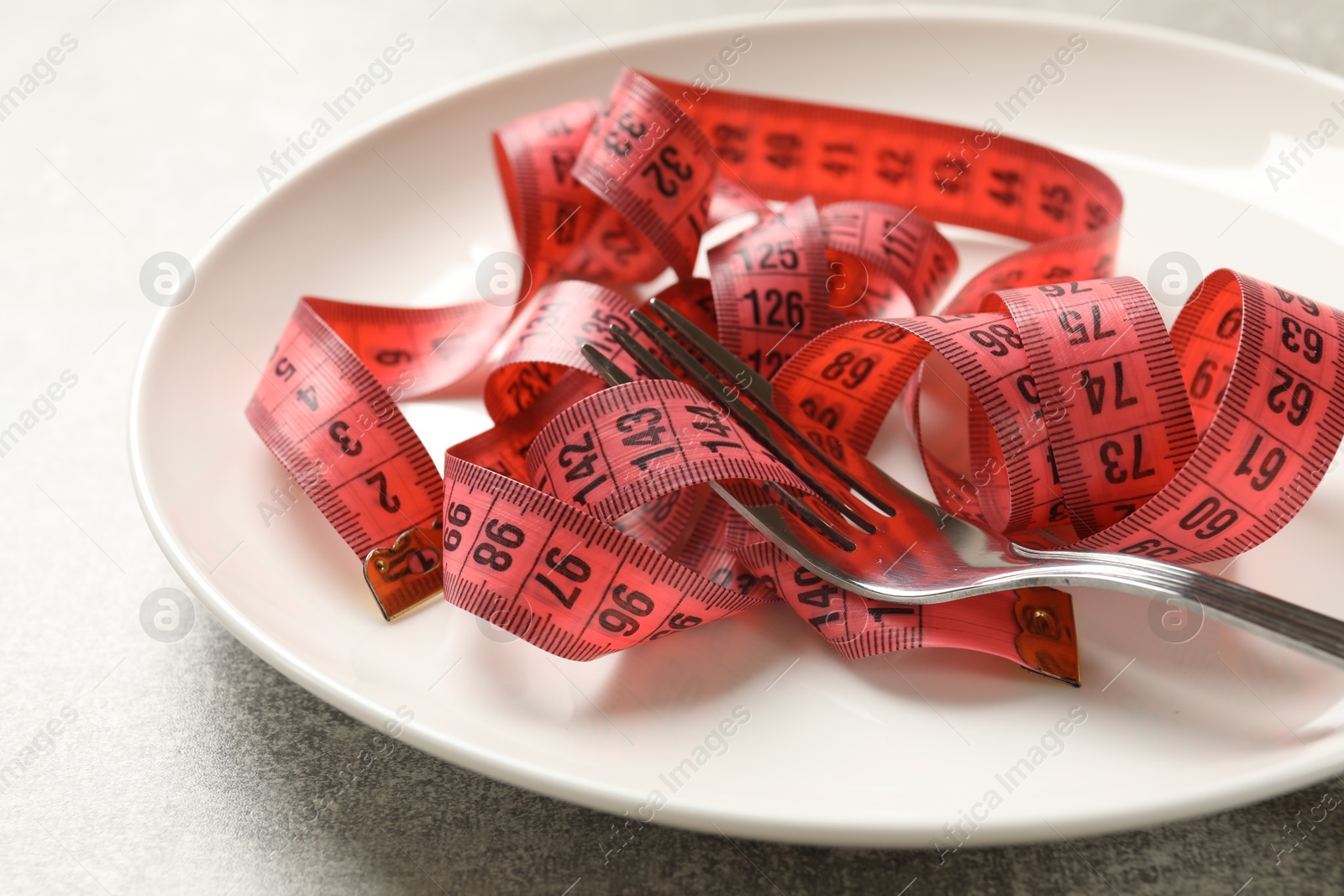 Photo of Bulimia. Fork, plate and measuring tape on grey table, closeup