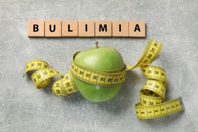 Photo of Squares with word Bulimia, green apple and measuring tape on grey table, flat lay