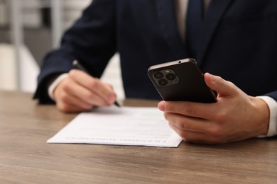 Photo of Man with smartphone signing licensing agreement document at wooden table indoors, closeup