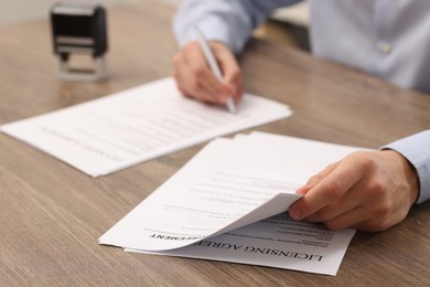 Photo of Man signing licensing agreement document at wooden table indoors, closeup