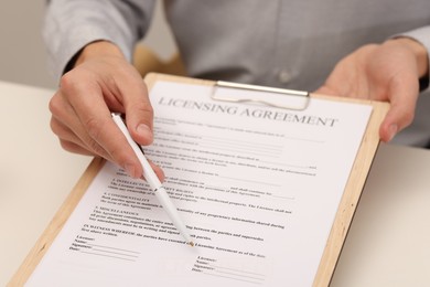 Photo of Man with pen pointing licensing agreement at document at white table indoors, closeup