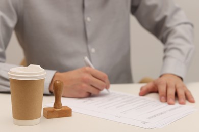 Photo of Man signing licensing agreement document at white table, focus on paper cup of drink and stamp