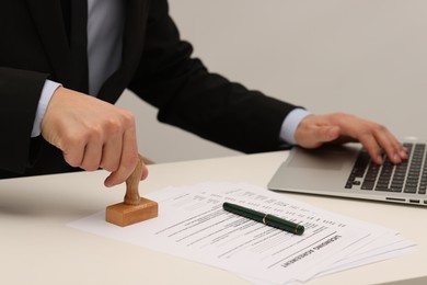 Photo of Man stamping licensing agreement document while working on laptop at white table indoors, closeup