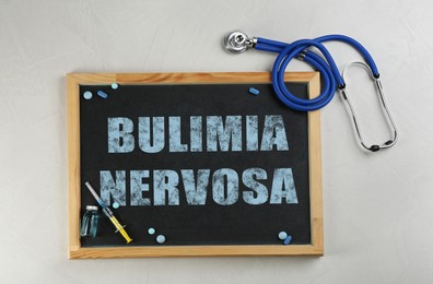 Image of Chalkboard with words Bulimia Nervosa, drugs and stethoscope on light background, flat lay