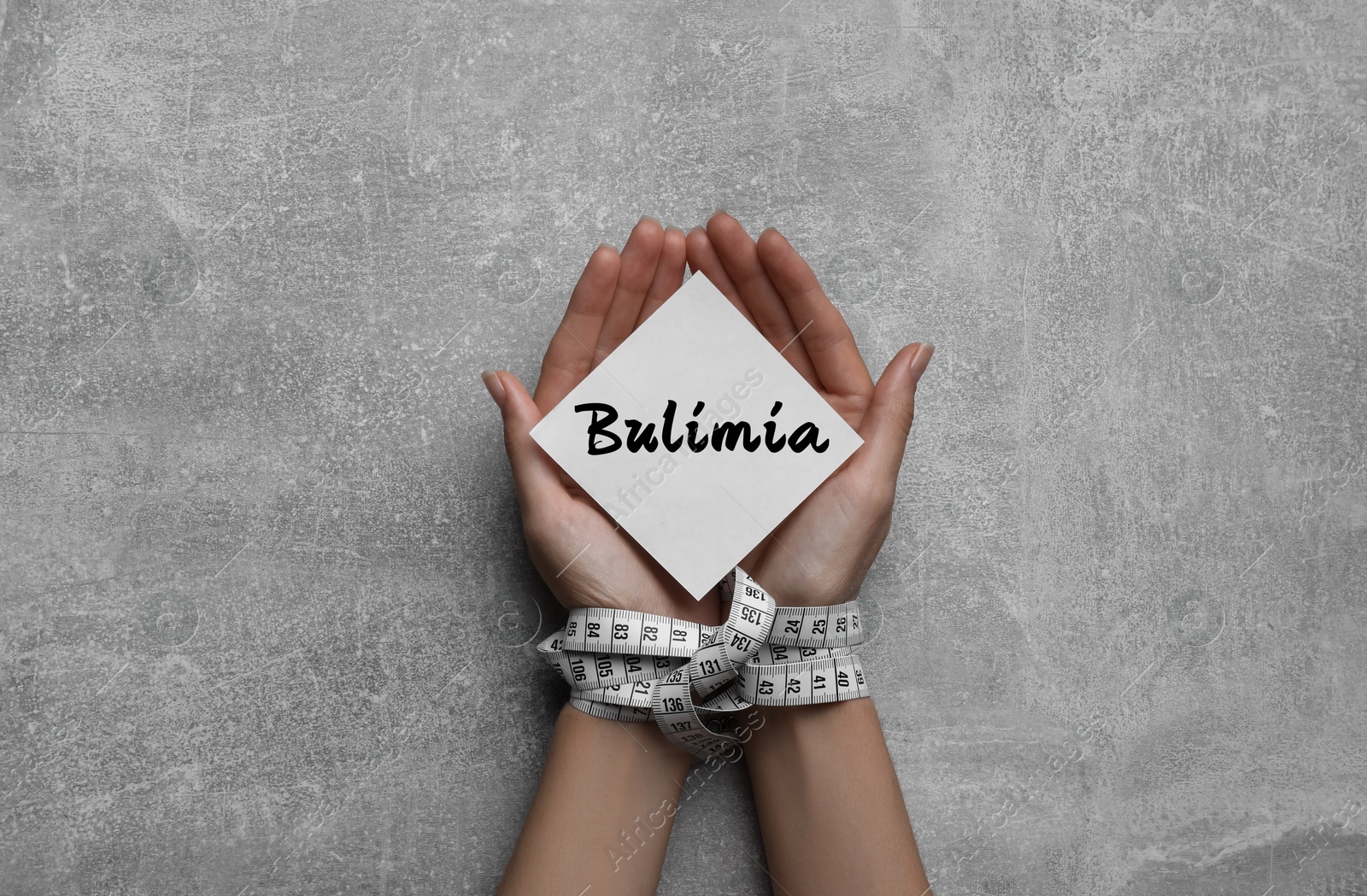 Image of Woman holding paper note with word Bulimia in hands tied with measuring tape on grey table, top view