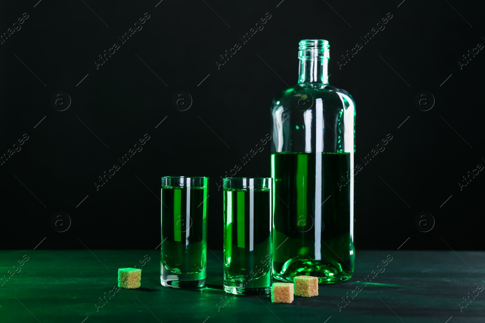 Photo of Absinthe in shot glasses, bottle and brown sugar on table against black background