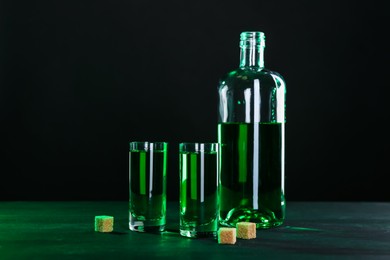 Photo of Absinthe in shot glasses, bottle and brown sugar on table against black background