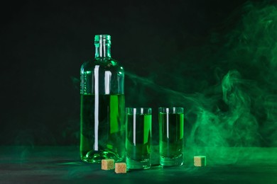 Photo of Absinthe in shot glasses, bottle and brown sugar on table against black background with smoke