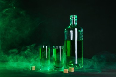 Photo of Absinthe in shot glasses, bottle and brown sugar on table against black background with smoke