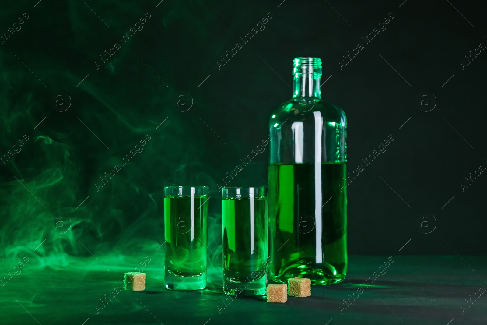 Photo of Absinthe in shot glasses, bottle and brown sugar on table against black background with smoke