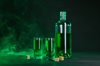 Photo of Absinthe in shot glasses, bottle and brown sugar on table against black background with smoke