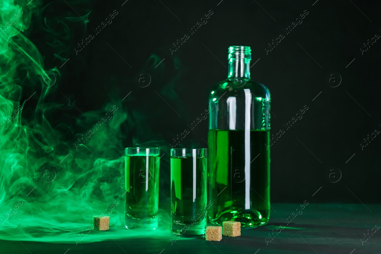 Photo of Absinthe in shot glasses, bottle and brown sugar on table against black background with smoke