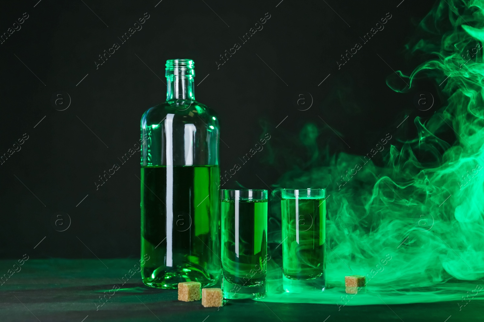 Photo of Absinthe in shot glasses, bottle and brown sugar on table against black background with smoke