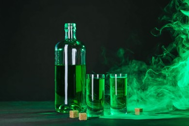 Photo of Absinthe in shot glasses, bottle and brown sugar on table against black background with smoke