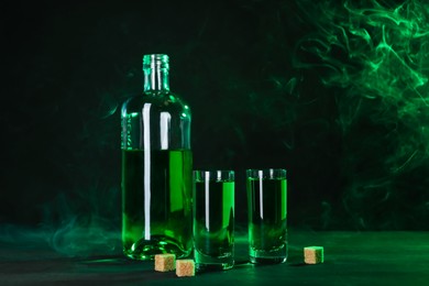 Photo of Absinthe in shot glasses, bottle and brown sugar on table against black background with smoke