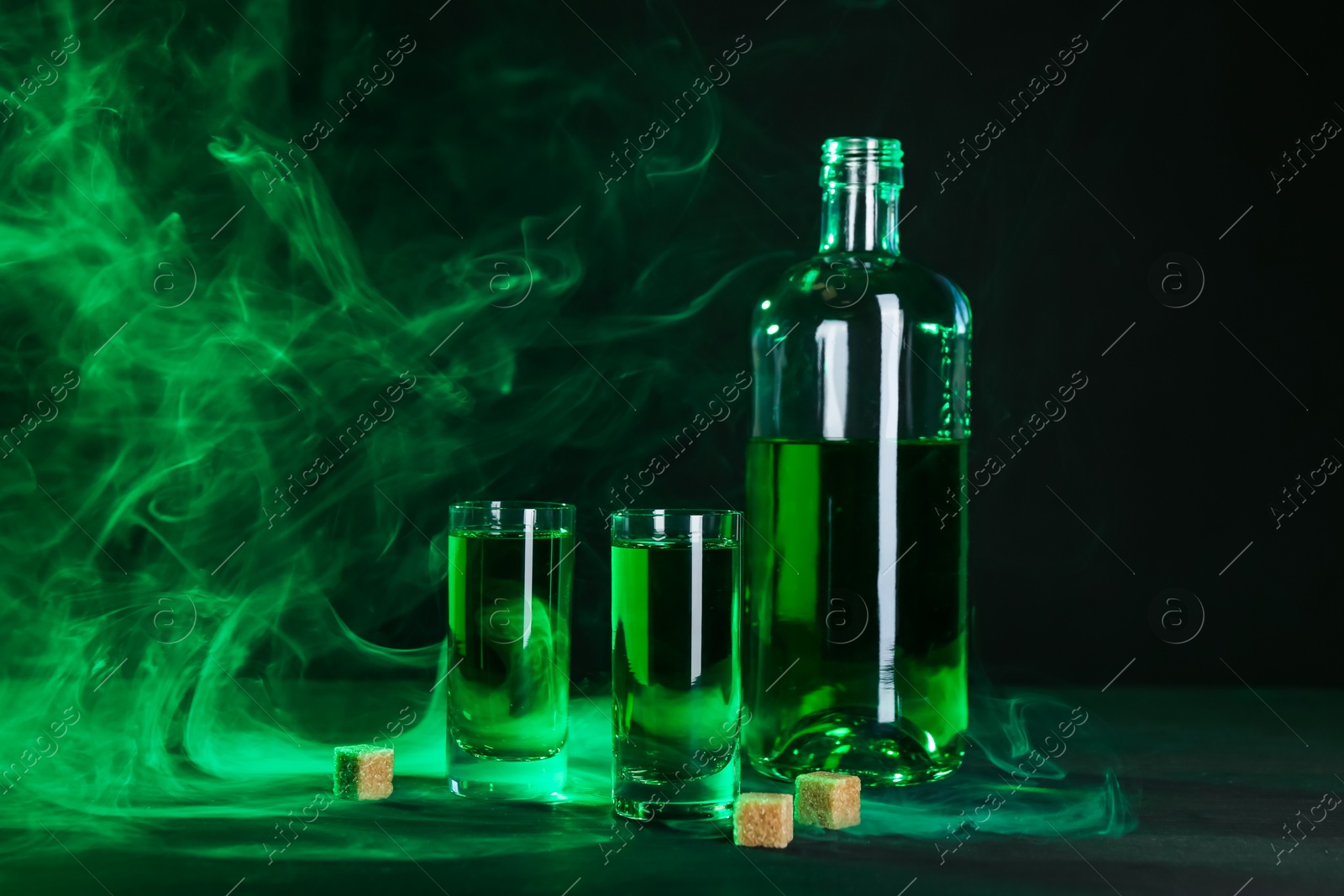 Photo of Absinthe in shot glasses, bottle and brown sugar on table against black background with smoke