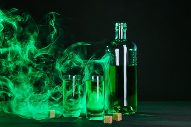 Photo of Absinthe in shot glasses, bottle and brown sugar on table against black background with smoke