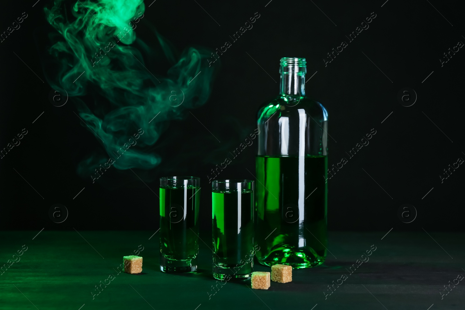 Photo of Absinthe in shot glasses, bottle and brown sugar on table against black background with smoke