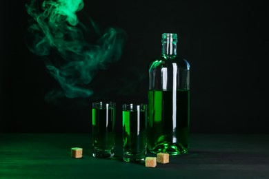 Photo of Absinthe in shot glasses, bottle and brown sugar on table against black background with smoke