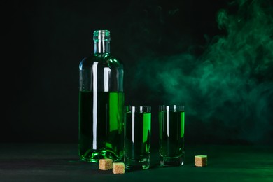 Photo of Absinthe in shot glasses, bottle and brown sugar on table against black background with smoke