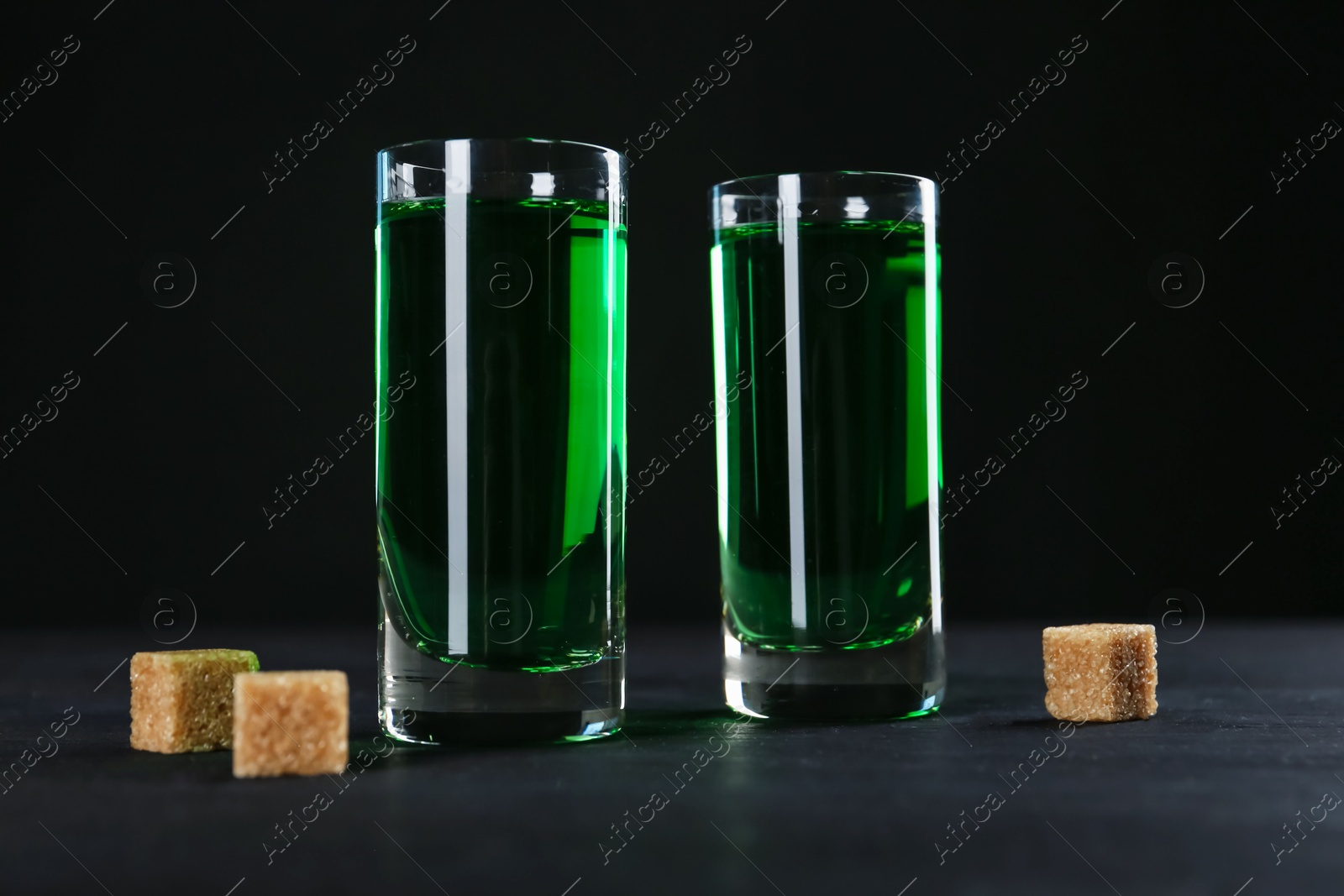 Photo of Absinthe in shot glasses and brown sugar on table against black background, closeup