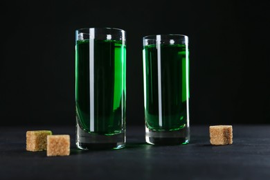 Photo of Absinthe in shot glasses and brown sugar on table against black background, closeup