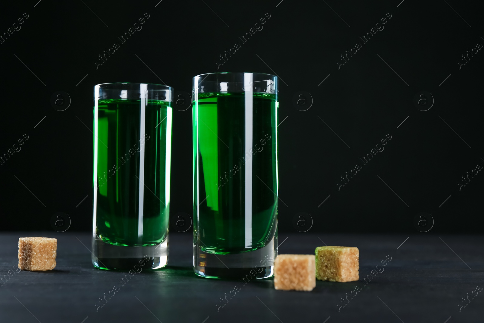 Photo of Absinthe in shot glasses and brown sugar on table against black background, closeup