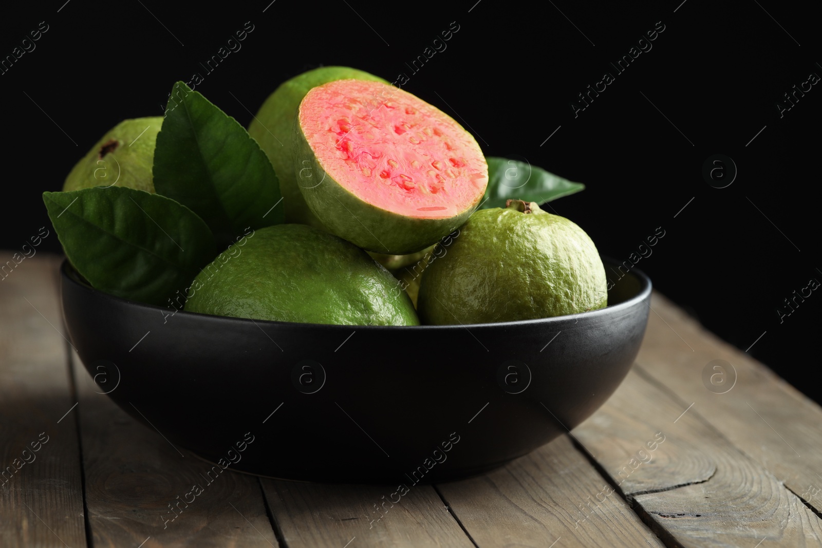 Photo of Fresh guava fruits in bowl on wooden table against black background, closeup