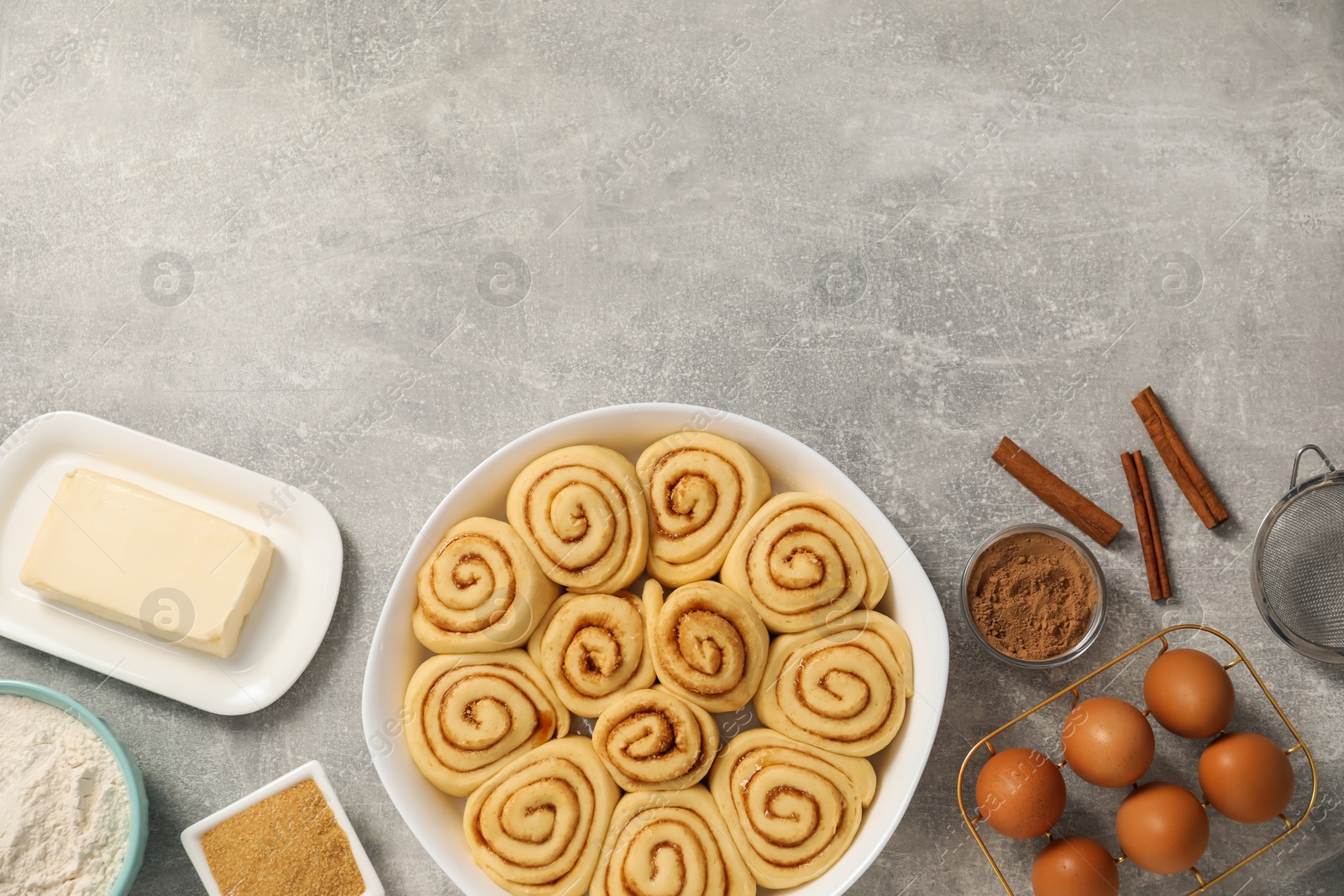 Photo of Raw cinnamon rolls in baking dish and different ingredients on grey table, flat lay. Space for text