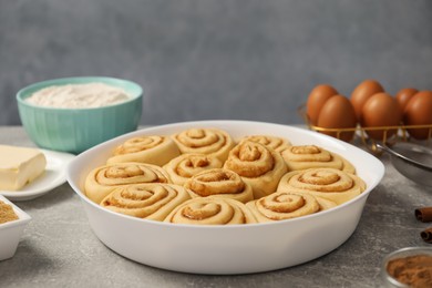 Raw cinnamon rolls in baking dish and different ingredients on grey table, closeup