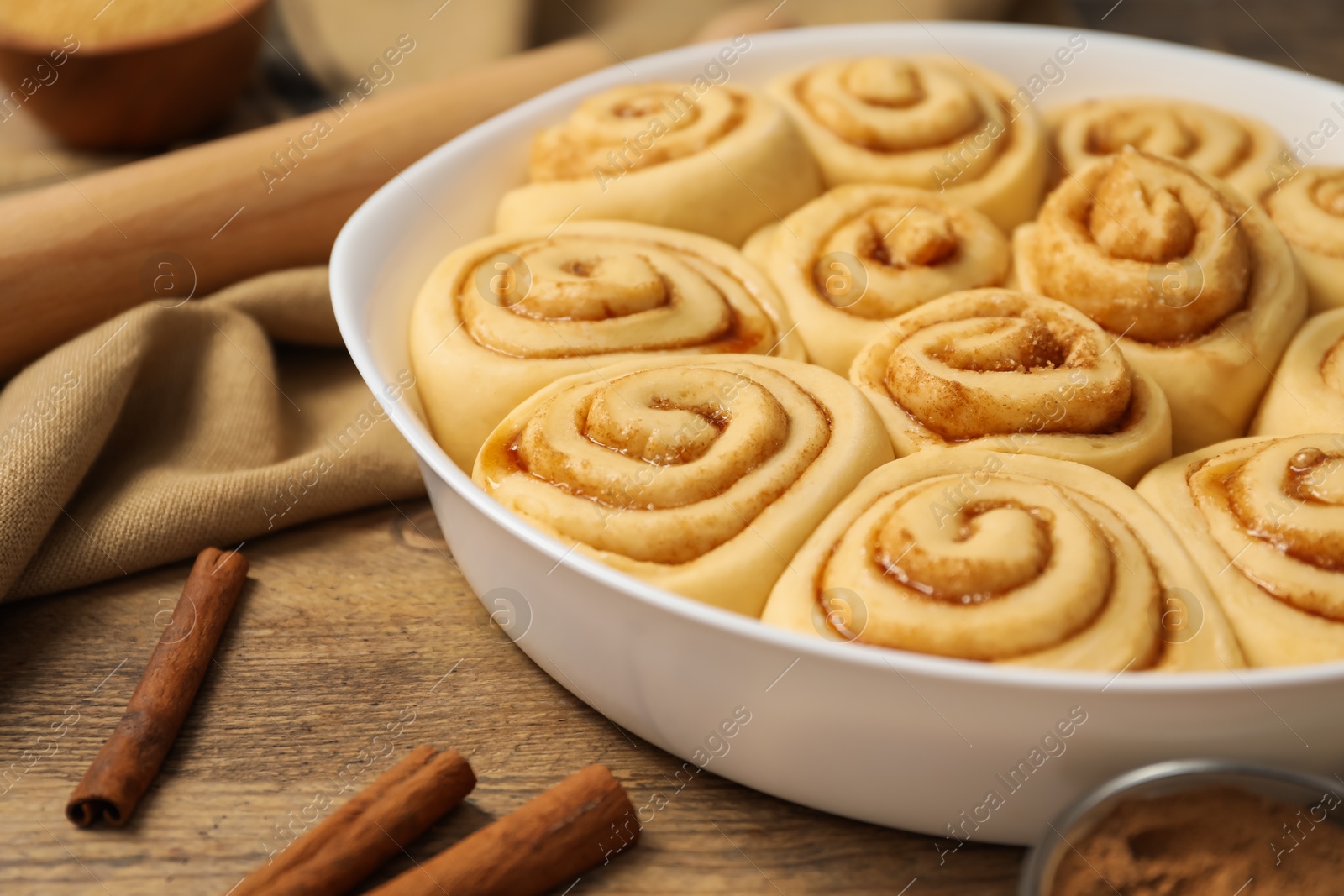 Photo of Raw cinnamon rolls in baking dish, sticks and cocoa powder on wooden table, closeup