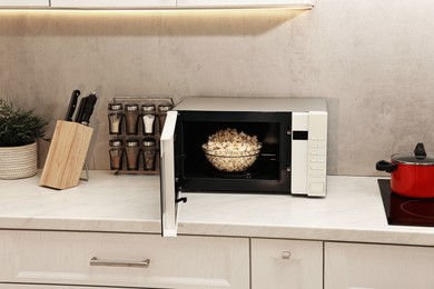 Photo of Open microwave oven with bowl of popcorn on white marble countertop in kitchen