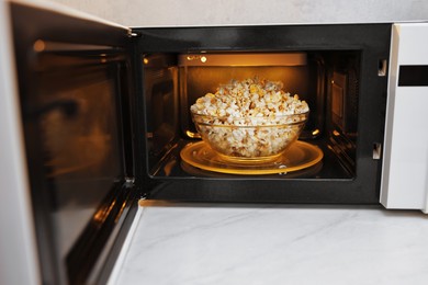 Photo of Open microwave oven with bowl of popcorn on white marble countertop in kitchen