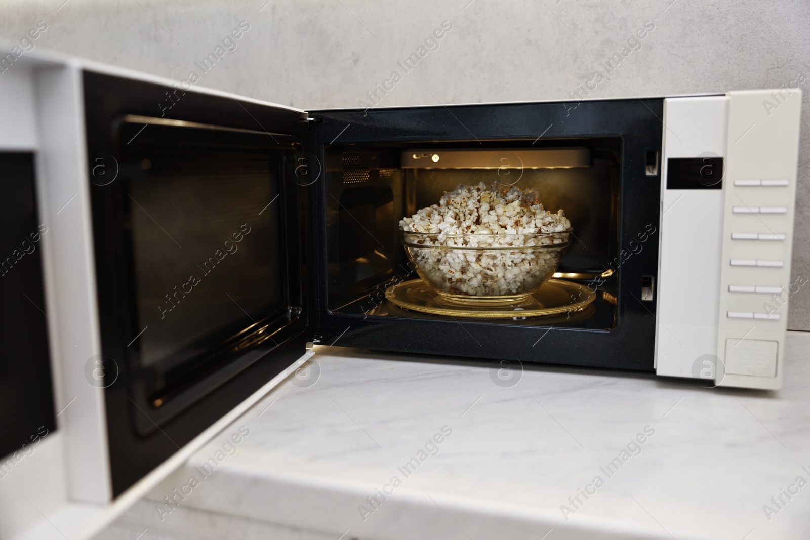 Photo of Open microwave oven with bowl of popcorn on white marble countertop in kitchen