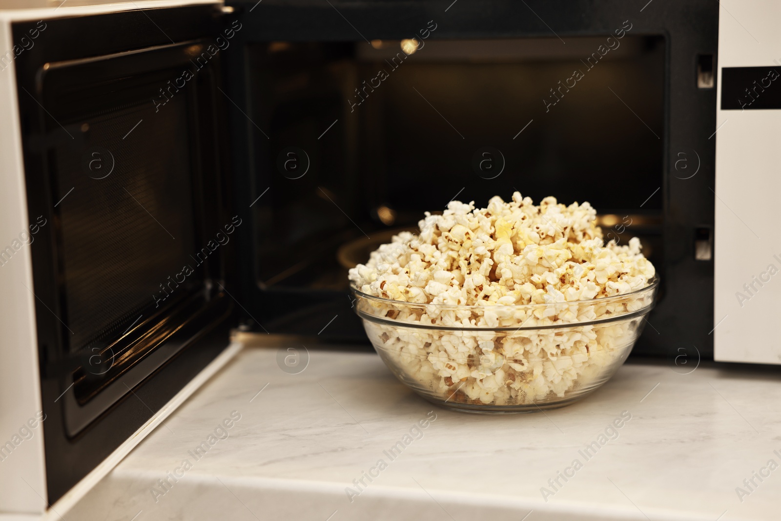 Photo of Tasty popcorn in bowl near microwave oven on white marble countertop indoors