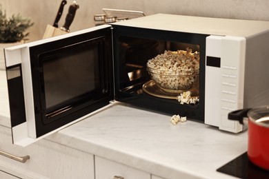 Photo of Open microwave oven with bowl of popcorn on white marble countertop in kitchen