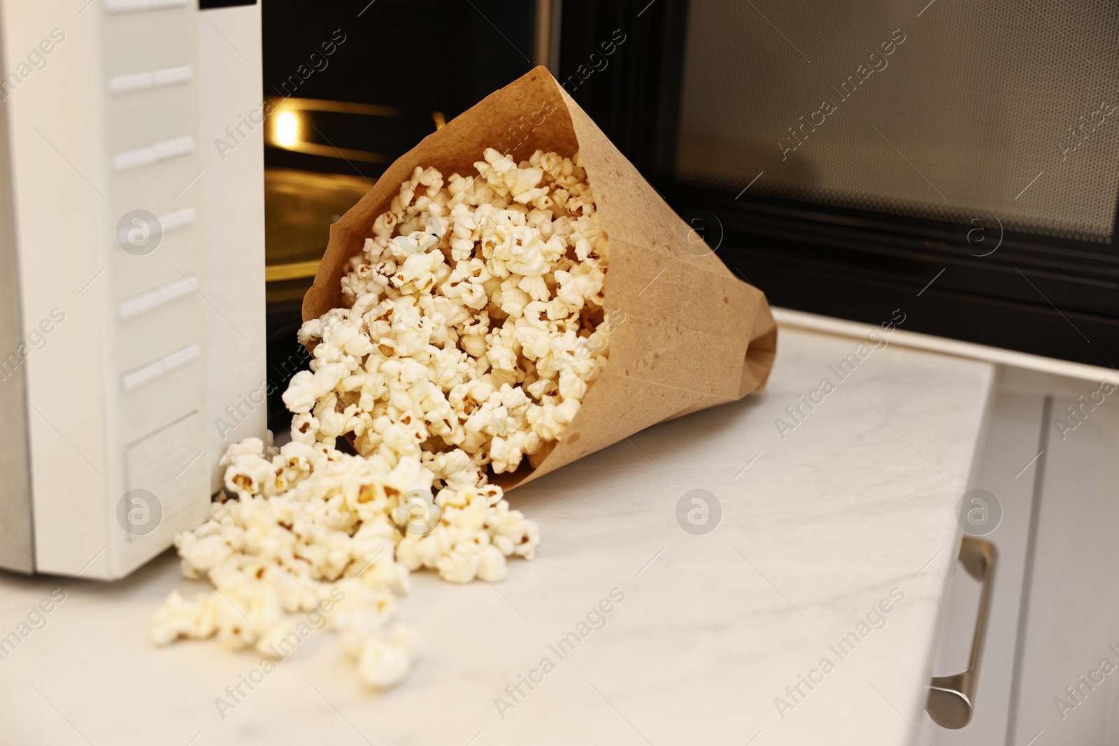 Photo of Tasty popcorn in paper bag near microwave oven on white marble countertop indoors, closeup