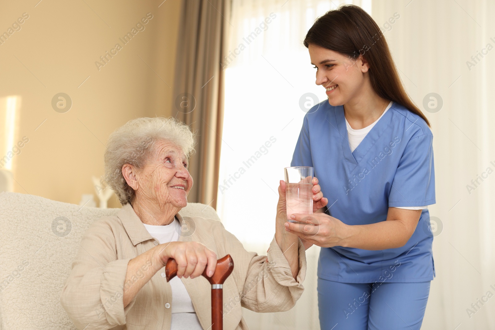 Photo of Healthcare worker giving elderly woman glass of water indoors