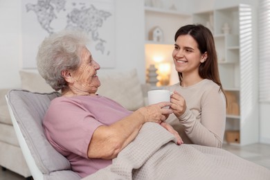 Photo of Granddaughter giving hot drink to her grandmother at home. Elderly care