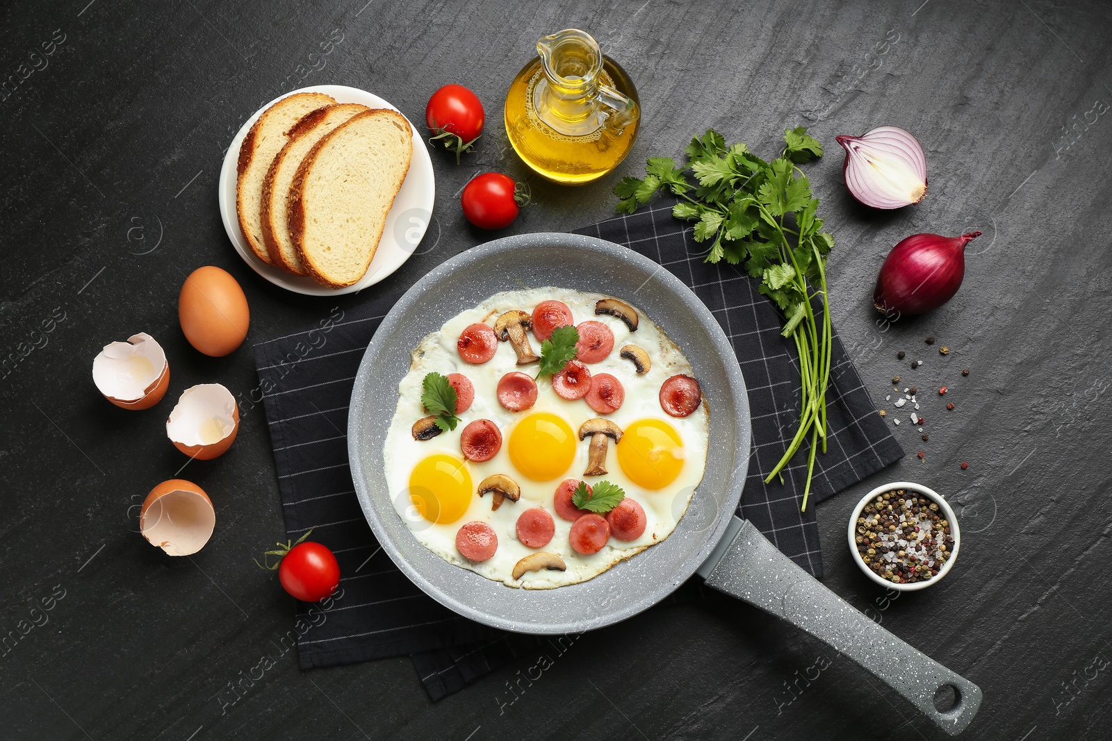 Photo of Tasty fried eggs with cut sausages and mushrooms in frying pan surrounded by ingredients on black table, flat lay